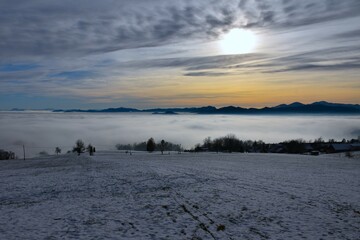 Snow covered field above fogg landscape of Gorenjska, Slovenia with hills behind