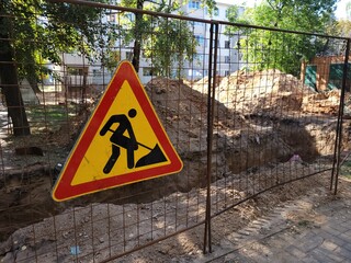 Construction site on city street is marked by metal barricade with triangular warning sign...