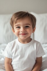 Fototapeta premium A cheerful young baby boy with short brown hair, wearing a plain white t-shirt mockup , smiles warmly at the camera while sitting on a bed with white bedding.