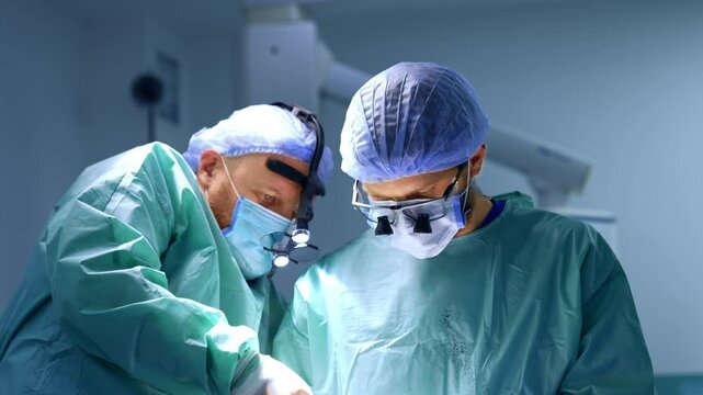 Bearded doctor uses a screw-driver at operation and passes it to the nurse. Male surgeons wearing robes stained with blood cooperate at surgery.