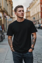 Fototapeta premium A young man with tattoos stands confidently on a city street, wearing a black t-shirt mockup and jeans. The background features urban architecture and a cobblestone road.
