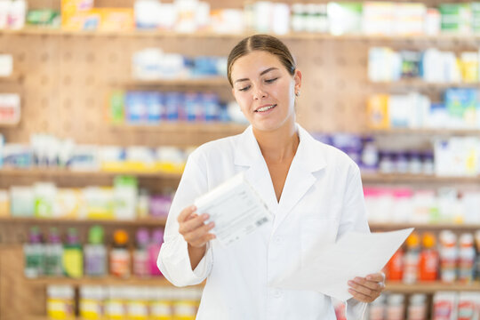 Positive young female pharmacist standing in chemist's shop holding box with medicament and a sheet of recipe