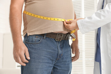 Nutritionist measuring overweight man's waist with tape in clinic, closeup