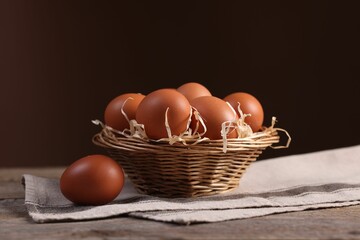Wicker basket with fresh eggs on wooden table against brown background, closeup