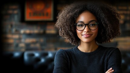 A woman with curly hair and glasses is smiling at the camera. She is wearing a black sweater and is standing in front of a brick wall