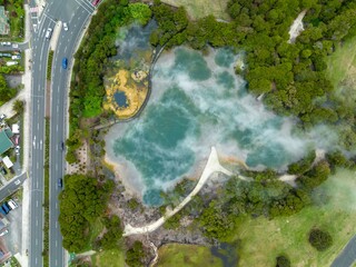 Aerial view of a geothermal pool, surrounded by lush greenery and a paved walkway. Steam rises from...