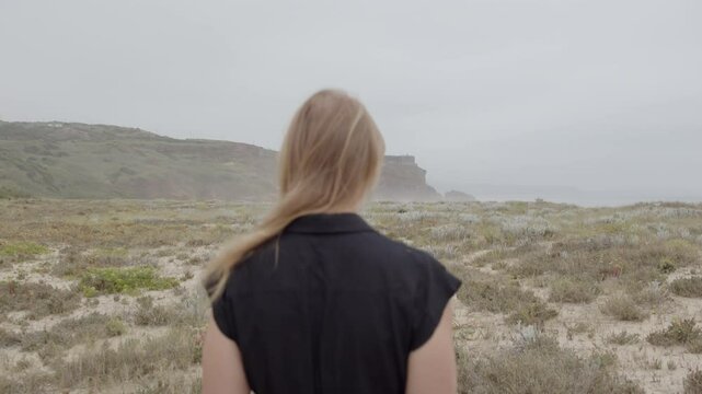 Blonde woman filmed from the back with no face visible wearing a black dress walking away from the camera towards the ocean in a desolate landscape