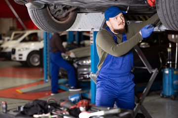 Young guy mechanic in uniform repairs underbody of car in car service station