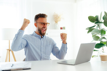 Naklejka premium Cheerful successful businessman with glasses in a blue shirt, raising his fists while looking at his laptop screen, celebrating a success while sitting in a bright modern office, smiling happily