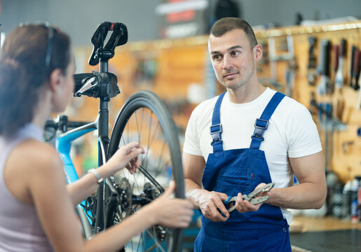 Professional confident bicycle technician with cassette pliers in hands examining bike wheel and discussing repair details with female client in workshop....