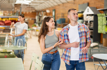 Positive friendly young adult couple, guy and girl, looking with interest for comfortable furniture for patio and garden of country house in home decor store