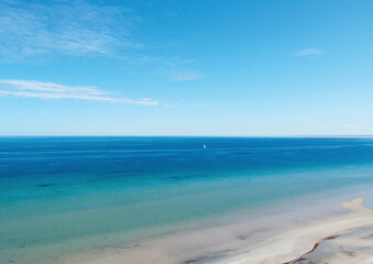 Fototapeta premium Aerial view of calm ocean waves under a vibrant blue sky with a distant sailboat