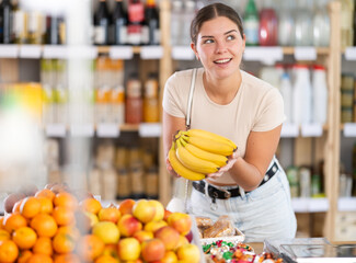Dark-haired girl buys bananas in store. She takes ripe fruits out of display case, inspects goods...