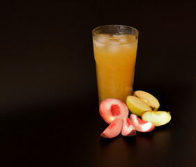 Tall glass of fruit juice mix with ice on black background, pieces of ripe peach and apple nearby.
