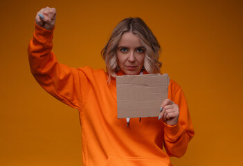 Woman in an orange hoodie,raising fist in the air while holding torn cardboard piece.Her serious...