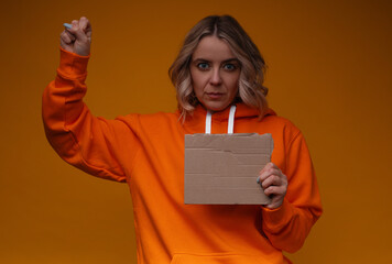 Naklejka premium A woman in an orange hoodie, holding a piece of torn cardboard in one hand and raising a fist with the other, set against a vibrant orange background. The image conveys empowerment and activism.