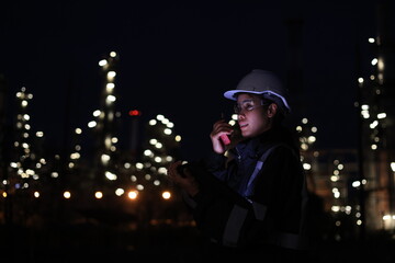 A female chemical worker in safety gear uses a digital tablet and walkie-talkie at a brightly lit refinery at night