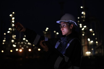 A female chemical worker in safety gear uses a digital tablet and walkie-talkie at a brightly lit refinery at night