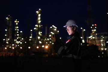 A female chemical worker in safety gear uses a digital tablet and walkie-talkie at a brightly lit refinery at night