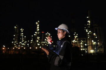 A female chemical worker in safety gear uses a digital tablet and walkie-talkie at a brightly lit refinery at night