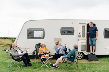 Group of older adults enjoying a day by a caravan. Relaxing outdoors, chatting, and reading. Casual gathering by caravan, sharing leisure time together. Happy mature friends on camping trip in nature.