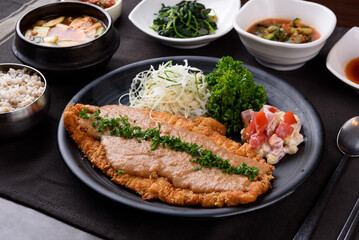 Pork cutlet and salad and parsley are placed on a black plate, and rice, soybean paste stew, vegetables, and kimchi are on the side.