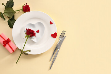 Beautiful table setting with red roses, gift box and hearts on yellow background. Valentine's Day celebration