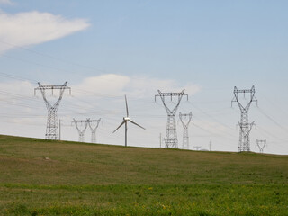 Wind turbine and power lines in a grassy field under a partly cloudy sky.  A landscape of renewable and traditional energy sources.