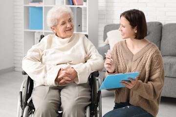 Fototapeta premium Young nurse with clipboard and senior woman in wheelchair at home