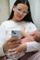 Beautiful young woman wearing white clothes is breastfeeding her baby at home, close-up back view. She is working online at the same time, remote work from home. 