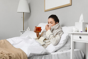 Ill young woman with lemon tea sneezing in bedroom