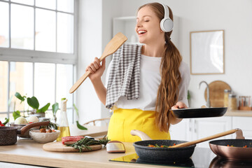 Young housewife in headphones with frying pan and spatula singing at home