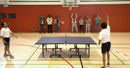 Playing table tennis, two men competing in school gymnasium with spectators watching - Powered by Adobe