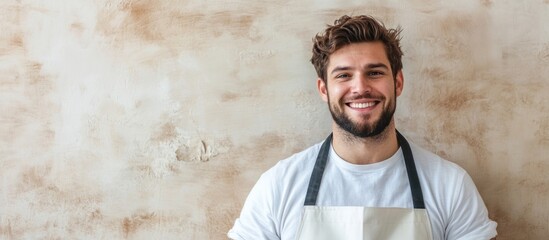 Fototapeta premium smiling man in white t-shirt and apron against textured beige wall with large empty space for text in bright lighting and cheerful atmosphere