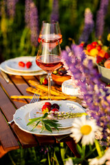 Two glasses of rose wine, flowers in a vase, silverware, fruits. Sunset, golden hour, atmospheric candlelight. Romantic table decor for a loving couple on blooming meadow with purple lupines