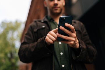 Cropped shot of young man standing on city street, using holding smartphone to check messages, browse internet, stay connected enjoying urban environment, selective focus. Concept of urban lifestyle.