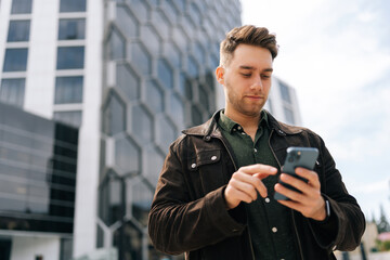 Low-angle portrait of focused successful business man using smartphone outside modern office center, confidently engaging in business communication on mobile app, looking to screen outdoors. © dikushin