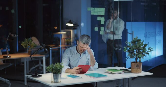 Couple of caucasian executives staying long hours in dark office. Tired adult businessman using tablet computer solving company problems negotiating meeting.