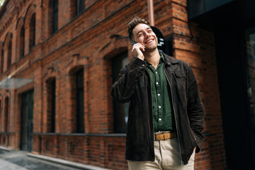 Naklejka premium Portrait of joyful young man happily talking on smartphone while walking near brick building, enjoying conversation and urban atmosphere. Concept of modern successful city lifestyle.