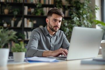 Focused young man typing on a laptop in a bright office, surrounded by greenery and bookshelves. Concept of productivity, work, and modern workspace. Ai generative