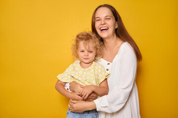 A mother holds her smiling daughter in a cozy room, both dressed casually. The vibrant yellow background adds warmth as they enjoy a happy moment together