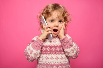 A young child with curly hair is holding a phone to her ear, eyes wide open in excitement, with her hands on her cheeks, surrounded by a bright pink background