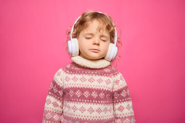 A child with curly hair listens to music, wearing large white headphones, with eyes closed and a serene expression, set against a bright pink backdrop