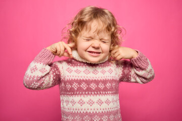 A young child with curly hair wears a pink patterned sweater and scrunches their face while...