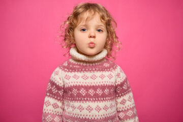 A young child with curly hair playfully pouts while wearing a patterned sweater. The bright pink backdrop creates a lively and fun atmosphere, showcasing the child's joyful personality