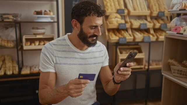 Young man in a bakery shop holding a smartphone and credit card, looking at his phone with various baked goods displayed on shelves in the background