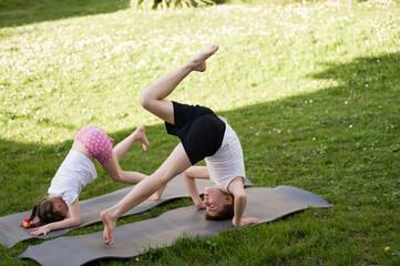 Two children playfully practice yoga poses on exercise mats outdoors. The vibrant green grass forms a natural backdrop to their active fun in the sunshine.