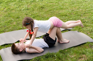 Two children, a girl and a boy, engage in playful acro-yoga on exercise mats outside. They are...