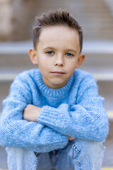 A confident young boy, dressed in a cozy blue sweater, sits outside with a serious expression, making him an ideal subject for family and childrens fashion themes that emphasize style and comfort