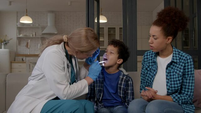 Positive female ENT doctor in uniform examining mouth and throat of sick teenage black boy using tongue depressor and flashlight, finding out cause of illness while visiting black family at home.